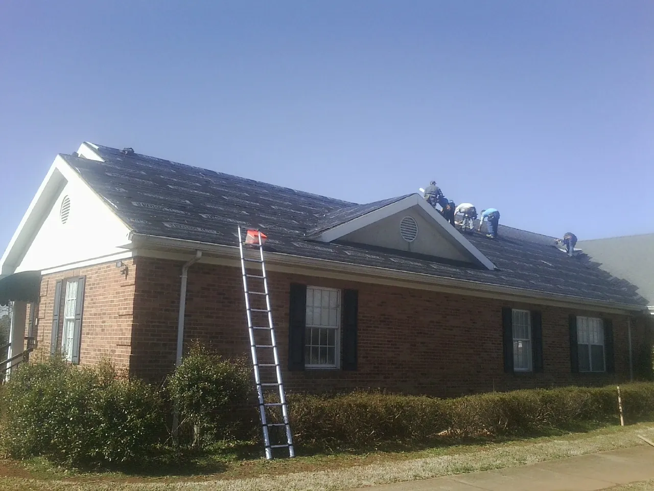 Crew tearing shingles off a brick ranch roof with a ladder set — Metal Roof Repair work by our crew in White Lake